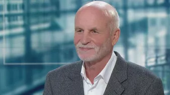 Homme âgé avec cheveux gris, souriant, portant un costume sobre sur fond flou.