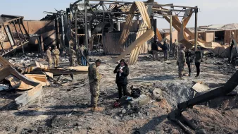 L'image montre une scène de destruction, probablement après un bombardement ou un incendie. On peut voir des débris et des structures en ruines, avec des panneaux de bois fissurés. Plusieurs militaires et civils se trouvent sur le site, certains en train de prendre des notes ou d'observer les dégâts. L'atmosphère semble grave, soulignant les conséquences d'un conflit. Le ciel est clair, contrastant avec le désastre au sol.