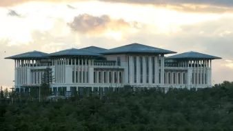 L'image montre un bâtiment impressionnant, probablement un complexe gouvernemental ou administratif. Il possède une architecture moderne, avec plusieurs niveaux et des toits en pente. Les murs sont principalement en blanc, avec de grandes fenêtres, ce qui laisse penser à un design spacieux et lumineux. Le bâtiment est entouré d'un paysage verdoyant, ce qui ajoute à son aspect majestueux. Le ciel en arrière-plan est nuageux, ce qui crée une ambiance dramatique.