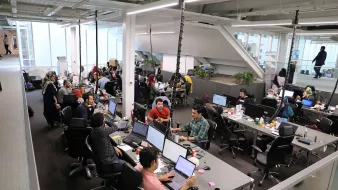 L'image montre un espace de bureau moderne et dynamique. Il y a plusieurs personnes qui travaillent à des bureaux équipés d'ordinateurs. L'environnement semble collaboratif, avec des employés assis autour de tables, certains en train de discuter, tandis que d'autres se concentrent sur leur travail. Les bureaux sont bien éclairés et décorés avec des plantes. On peut voir des objets personnels et des collations éparpillés sur les tables, ce qui donne une atmosphère informelle et conviviale.