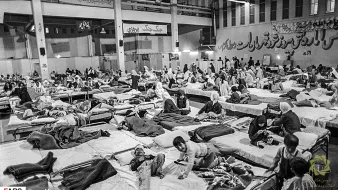 L'image montre un grand espace intérieur aménagé en salle de soins, avec de nombreux lits occupés par des personnes. On peut voir des patients allongés sur des matelas, certains semblant recevoir des soins médicaux. L'atmosphère est chargée et il y a un sentiment d'urgence, avec une présence significative de personnel soignant et de familles. Les murs portent des inscriptions en persan, ajoutant à l'authenticité du lieu.