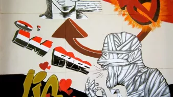 L'image représente une œuvre de street art colorée. On y voit un personnage avec des lunettes de soleil et une expression déterminée, entouré de textes. Au-dessus, une flèche pointe vers le haut avec l'inscription "I'M FREE" (je suis libre) en lettres stylisées. À droite, un autre personnage est représenté, désigné comme étant bandé, avec des gestes qui semblent exprimer une forte émotion. Le fond combine des couleurs vives comme le rouge et le jaune, avec des cœurs dessinés au bas, ce qui ajoute une touche plus douce à l'ensemble. L'œuvre montre un contraste entre la liberté et une certaine souffrance, évoquant des thèmes profonds.