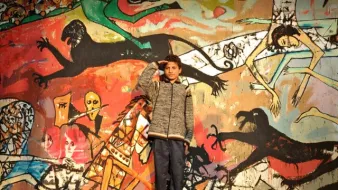 L'image montre un jeune garçon en train de faire un salut militaire devant un mur coloré, sur lequel sont peintes diverses figures stylisées et abstraites. Le mur présente des scènes dynamiques, avec des motifs de monstres et d'êtres humains, créant un contraste frappant avec le garçon qui porte un manteau gris et un pantalon sombre. Son expression et sa posture semblent témoigner de confiance ou de détermination. Les couleurs vives et les formes exagérées de la fresque attirent l'œil et ajoutent une dimension artistique à la scène.