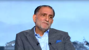 Homme en costume gris, assis, avec une expression pensante, arrière-plan flou.