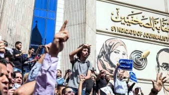 L'image montre une scène de manifestation devant un bâtiment, probablement le Syndicat des journalistes. De nombreuses personnes sont rassemblées, levant les bras en signe de protestation. Certains tiennent des pancartes ou des banderoles, tandis qu'une personne debout sur une foule semble s'adresser aux manifestants. L'arrière-plan présente une grande fresque représentant des figures, ajoutant à l'impact visuel de la scène. L'atmosphère est celle d'un rassemblement engagé et passionné.