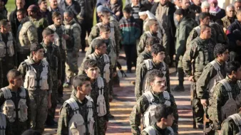 L'image montre un groupe de soldats en formation, portant des uniformes militaires et des équipements. Ils sont alignés, en regardant droit devant eux, et semblent être en train de participer à une cérémonie ou à un événement officiel. En arrière-plan, on peut observer une foule de personnes, probablement des civils, qui les regardent. L'atmosphère semble sérieuse et solennelle.