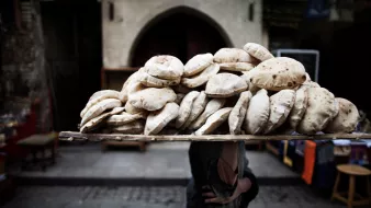 L'image montre une personne portant une grande pile de pains sur un plateau placé sur sa tête. Les pains, ronds et légèrement plats, semblent fraîchement cuits et sont empilés de manière ordonnée. En arrière-plan, on peut apercevoir des éléments typiques d'un marché, avec des structures architecturales anciennes et un éclairage ambiant qui donne une atmosphère vivante à la scène. L'accent est mis sur le transport traditionnel des marchandises en milieu urbain.