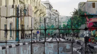 L'image montre un mur de blocs de béton, sur lequel sont peints des paysages urbains. La peinture représente une rue avec des arbres, des piétons et des cyclistes. On peut voir des silhouettes de personnes et des éléments de la ville, comme des lampadaires. Ce contraste entre le mur brut et l'art coloré apporte une touche dynamique à l'environnement urbain. Le ciel est nuageux, ajoutant une atmosphère un peu dramatique à la scène.