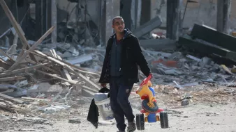 L'image montre un homme marchant dans un paysage dévasté. Il porte un seau de peinture dans sa main gauche et un jouet pour enfant, un petit véhicule à roues, dans sa main droite. L'environnement autour de lui est marqué par des débris et des ruines, suggérant un endroit qui a subi des destructions importantes. L'homme a l'air déterminé malgré la situation difficile.