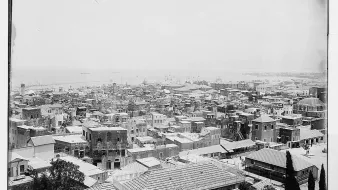 L'image présente une vue panoramique d'une ville côtière. On peut voir de nombreux bâtiments aux toits en tôle, ainsi que des structures en maçonnerie. La mer est visible à l'arrière-plan, avec quelques bateaux amarrés. Les rues semblent étroites et bien dessinées, et il y a des arbres éparpillés dans la zone. L'ensemble donne une impression de densité urbaine et de vie maritime. La photographie semble ancienne, capturant l'architecture et l'ambiance d'une époque révolue.