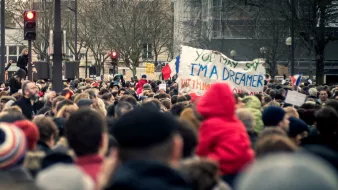L'image montre une grande foule rassemblée, probablement pour une manifestation ou une marche. On peut voir des personnes de tous âges, certaines portant des écharpes ou des bonnets, tandis que d'autres sont vêtues de manteaux. Au centre, il y a une pancarte sur laquelle est écrit : "YOU MAY SAY I'M A DREAMER BUT I'M NOT THE ONLY ONE". Des drapeaux français sont visibles, ce qui suggère un contexte patriotique ou une revendication sociale. L'atmosphère semble être à la fois solide et engagée.