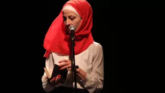 L'image montre une femme debout devant un microphone sur scène. Elle porte un hijab rouge vif et une blouse blanche. Elle tient un petit livre dans ses mains, semblant s'apprêter à lire ou à partager un texte. L'éclairage est sombre, mettant en valeur son visage et l'objet qu'elle tient. L'atmosphère semble intime et concentrée.