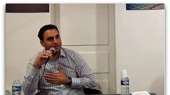 L'image montre un homme assis à une table, tenant un microphone. Il porte une chemise à rayures et semble s'exprimer avec conviction. Sur la table, il y a une bouteille d'eau et un gobelet, ainsi que des documents visibles. L'arrière-plan montre des affiches ou des œuvres, mais les détails ne sont pas clairement discernables.