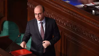 L'image montre un homme en costume sombre, portant des lunettes, se tenant près d'un pupitre dans une assemblée ou un parlement. Il semble être en train de s'adresser à l'assemblée ou de faire une déclaration informative, avec un dossier rouge posé sur le pupitre. Le décor est constitué de boiseries et de sièges verts, typiques d'un environnement institutionnel.