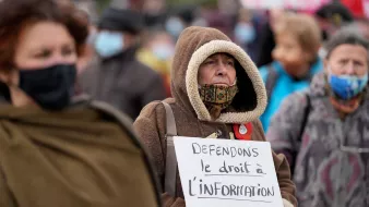 L'image montre un groupe de personnes rassemblées lors d'une manifestation. Au premier plan, une femme âgée porte une grande capuche et un masque, tenant une pancarte sur laquelle il est écrit : "Défendons le droit à l'information". Les autres manifestants sont flous en arrière-plan, mais on peut voir qu'ils portent également des masques. L'atmosphère semble sérieuse et engagée, avec des personnes qui militent pour leurs droits.