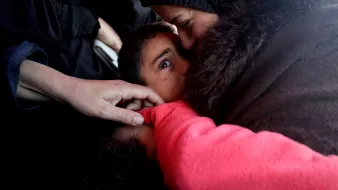 L'image montre un moment touchant entre un enfant et un adulte. L'adulte semble embrasser l'enfant, qui a l'air attentionné et gaze directement vers l'objectif. On peut aussi voir quelques mains, une d'elles touchant l'épaule de l'enfant. L'ambiance est empreinte d'émotion et de tendresse. Les vêtements des figures suggèrent un environnement austère.