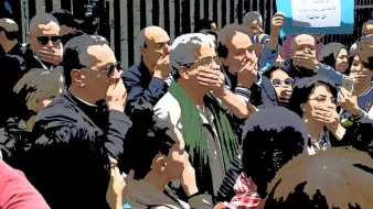 L'image montre un groupe de personnes réunies dans un cadre extérieur. Beaucoup d'entre elles se tiennent avec la main sur la bouche, semblant exprimer un silence ou protester. En arrière-plan, on aperçoit un panneau qui semble contenir un message. L'ambiance générale semble sérieuse et engagée, suggérant une forme de mobilisation ou de manifestation. Les participants semblent concentrés et solidaires.