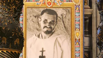 L'image montre un intérieur majestueux, probablement dans une église ou une cathédrale, avec des colonnes ornées de motifs dorés et des détails complexes. Au centre, il y a un grand portrait en noir et blanc d'un homme, qui semble vêtu d'une robe religieuse, encadré de couleurs vives et de décorations. Ce cadre attire l'œil et contraste avec la richesse des ornements de l'architecture environnante. Les murs et les éléments de la pièce évoquent une atmosphère sacrée et historique.