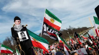 L'image montre une grande manifestation, probablement en faveur de la liberté ou des droits de l'homme. Au premier plan, une marionnette représentant un homme âgé est portée par un manifestant, tandis que d'autres personnes tiennent des drapeaux iraniens et des pancartes. Les participants semblent unis et déterminés, exprimant leurs revendications. L'ambiance est dynamique, avec une foule nombreuse et des symboles visuels forts liés à leur cause.