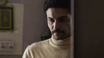 L'image montre un homme avec des cheveux bouclés et une moustache, portant un pull à col roulé de couleur claire. Il semble pensif, regardant vers le bas, avec une expression de réflexion ou de sérieux. En arrière-plan, on aperçoit des éléments de décoration intérieure, mais les détails sont flous. L'atmosphère de l'image dégage une certaine intimité ou mélancolie.