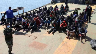 L'image montre un groupe de personnes assises sur le pont d'un bateau. Elles semblent être des migrants, plusieurs portant des vêtements chauds et des masques. À l'arrière, une personne en uniforme militaire observe la scène, tandis qu'un autre individu est vêtu d'un gilet portant un logo d'une organisation humanitaire. L'environnement semble être une zone portuaire, avec de l'eau visible à l'arrière-plan. L'atmosphère de l'image évoque une situation de contrôle ou d'assistance humanitaire.