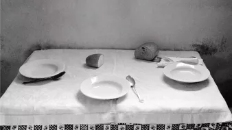 L'image montre une table couverte d'une nappe blanche, où se trouvent deux assiettes blanches, une cuillère et un couteau. Un petit pain est positionné sur un côté et un verre est renversé au centre de la table. L'ambiance générale est sobre et monochrome, ce qui donne une impression de simplicité et de calme. Les murs derrière la table sont légèrement ombragés, ajoutant un effet de profondeur à la composition.