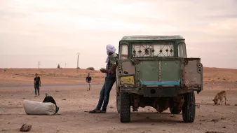 L'image montre une scène en plein air dans un paysage désertique. On peut voir un ancien véhicule utilitaire, avec une carrosserie usée et des fenêtres cassées. Un homme, partiellement visible, se tient à côté du véhicule, portant un turban. En arrière-plan, il y a d'autres personnes, ainsi qu'un chien qui déambule. Le ciel est nuageux, et l'atmosphère semble calme, mais aussi un peu désolée, avec des dunes de sable à l'horizon.