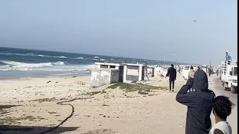 L'image montre une scène côtière avec des vagues de mer s'écrasant sur le rivage. Au premier plan, plusieurs personnes regardent vers l'horizon, peut-être fascinées par quelque chose dans le ciel. On peut également voir des véhicules stationnés sur le côté. L'ambiance semble calme, avec un ciel dégagé et une légère brise. La plage est principalement sablonneuse, et quelques éléments tels que des algues sont visibles sur le sol.