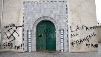 L'image montre un mur extérieur d'une mosquée, orné de carreaux colorés. Sur le mur, il y a des graffitis qui incluent un symbole controversé, représentant une swastika, ainsi que des phrases telles que "White power" et "La France aux Français." La porte d'entrée est en bois, avec des détails architecturaux typiques. L'ensemble de la scène évoque un message de haine et de division.