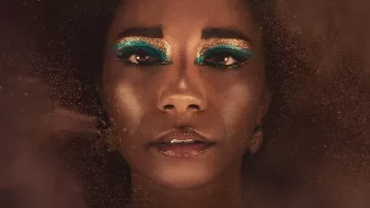 L'image montre une personne au visage ornée, avec un maquillage très artistique mettant en avant des yeux accentués par une ombre à paupières bleu vif. La personne porte une coiffe dorée en forme de serpents, ajoutant à l'aspect majestueux de l'ensemble. Le fond est flou et semble composé de particules dorées, créant une atmosphère mystique et intrigante. L'expression du visage est à la fois sereine et mystérieuse.