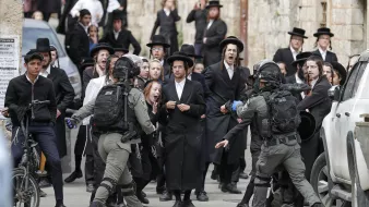 L'image montre une scène animée dans une rue, où un groupe de personnes, principalement habillées en noir, semble manifester ou réagir à une situation tendue. On peut voir des hommes en habits traditionnels, portant des chapeaux noirs, ainsi que des adolescents sur des bicycles. Au centre, des membres des forces de l'ordre, en tenue tactique, interagissent avec la foule. Les expressions faciales des personnes dans la foule varient entre la surprise, l'inquiétude et l'agitation, créant une atmosphère de tensions. Les bâtiments en arrière-plan évoquent un contexte urbain, probablement dans une région où se concentrent des communautés religieuses.