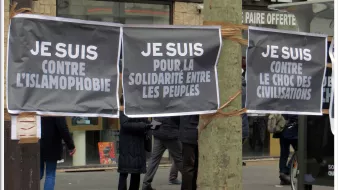 L'image montre plusieurs grandes affiches suspendues, avec des messages écrits en lettres blanches sur fond noir. Les slogans affichés expriment des positions contre l'islamophobie et en faveur de la solidarité entre les peuples. En arrière-plan, on peut apercevoir des personnes qui semblent participer à un rassemblement ou une manifestation. L'ambiance suggère une mobilisation citoyenne pour des causes sociales et politiques.
