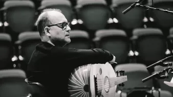 L'image montre un homme assis, tourné légèrement sur le côté. Il porte des lunettes et un pull noir. Dans ses bras, il tient un instrument de musique en forme de luth. L'arrière-plan est flou avec des rangées de sièges.