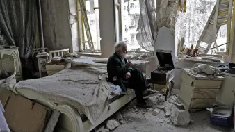 L'image montre un espace intérieur dévasté, probablement un appartement. Au centre, un homme âgé est assis sur un lit, tenant un objet à la main, peut-être une cigarette. L'environnement est marqué par des murs endommagés, des débris éparpillés et des fenêtres brisées, laissant voir l'extérieur. La lumière naturelle pénètre par les ouvertures, révélant l'état de désolation de la pièce. L'ambiance est sombre et mélancolique, illustrant les conséquences d'un conflit ou d'une catastrophe.