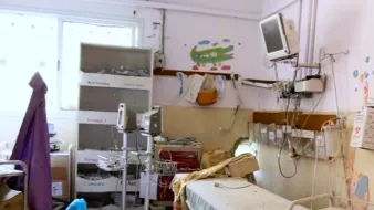 L'image montre une chambre d'hôpital qui a l'air un peu désordonnée et ancienne. On y voit un lit médical avec des draps, une étagère avec des boîtes, ainsi que du matériel médical. Un petit téléviseur est fixé au mur, et des illustrations colorées sont visibles sur les murs, ce qui ajoute une touche enfantine à l'environnement. La pièce semble nécessiter un peu de nettoyage et de modernisation.