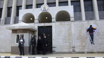 L'image montre un bâtiment arborant une façade en pierre. À l'avant, on voit une entrée avec des portes en fer forgé, encadrée par des arches. À gauche, un homme en costard consulte un document, tandis que deux autres hommes, également en costume, discutent à l'entrée. Un homme est sur une échelle, en train de fixer un panneau sur le mur. En arrière-plan, on distingue un poste de sécurité, suggérant que c'est un site administratif ou gouvernemental. Le sol est pavé en noir et blanc, ajoutant un contraste au décor.