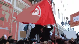 L'image représente une scène de manifestation. On peut voir un groupe de personnes, dont certaines tiennent des drapeaux et des pancartes. Un drapeau tunisien est clairement visible, symbolisant l'identité nationale et les revendications. Les manifestants semblent mobilisés et engagés, dans un environnement urbain avec des bâtiments en arrière-plan. L'atmosphère semble être celle d'une protestation pour des causes sociales ou politiques.