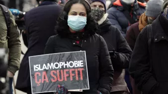 L'image montre une femme portant un masque, se tenant au milieu d'une foule. Elle tient une pancarte sur laquelle est écrit "ISLAMOPHOBIE ÇA SUFFIT". Les personnes autour d'elle semblent également s'engager dans une manifestation, témoignant d'un soutien à la cause qu'elle défend. L'atmosphère est sérieuse et engagée, reflétant des préoccupations sociales.