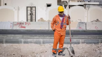 L'image montre un homme qui travaille sur un chantier de construction. Il porte un uniforme orange, ainsi qu'un casque de sécurité jaune. Il semble tenir une pelle, regardant vers le sol avec une posture réfléchie. En arrière-plan, on peut voir des murs de construction et des graffitis sur des surfaces environnantes. Le site semble en cours de développement, avec des matériaux de construction dispersés au sol.