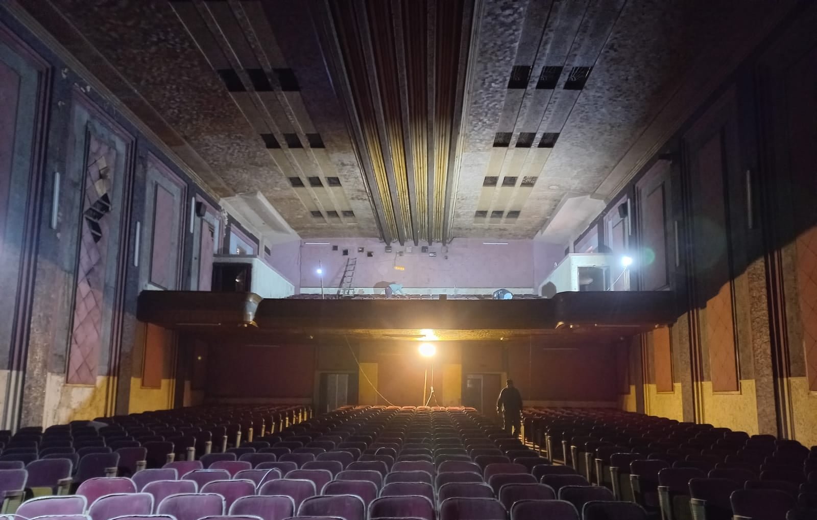 L'immagine mostra l'interno di un teatro abbandonato. Si possono vedere file di poltrone di velluto viola, di fronte a un palcoscenico che sembra vuoto. La luce proveniente da un proiettore evidenzia il pavimento e crea un'atmosfera suggestiva. Sul palcoscenico, ci sono alcune attrezzature, come scale, suggerendo che ci siano lavori in corso o recenti ispezioni. Le pareti del teatro mostrano segni di degrado, contribuendo a un senso di malinconia e nostalgia.