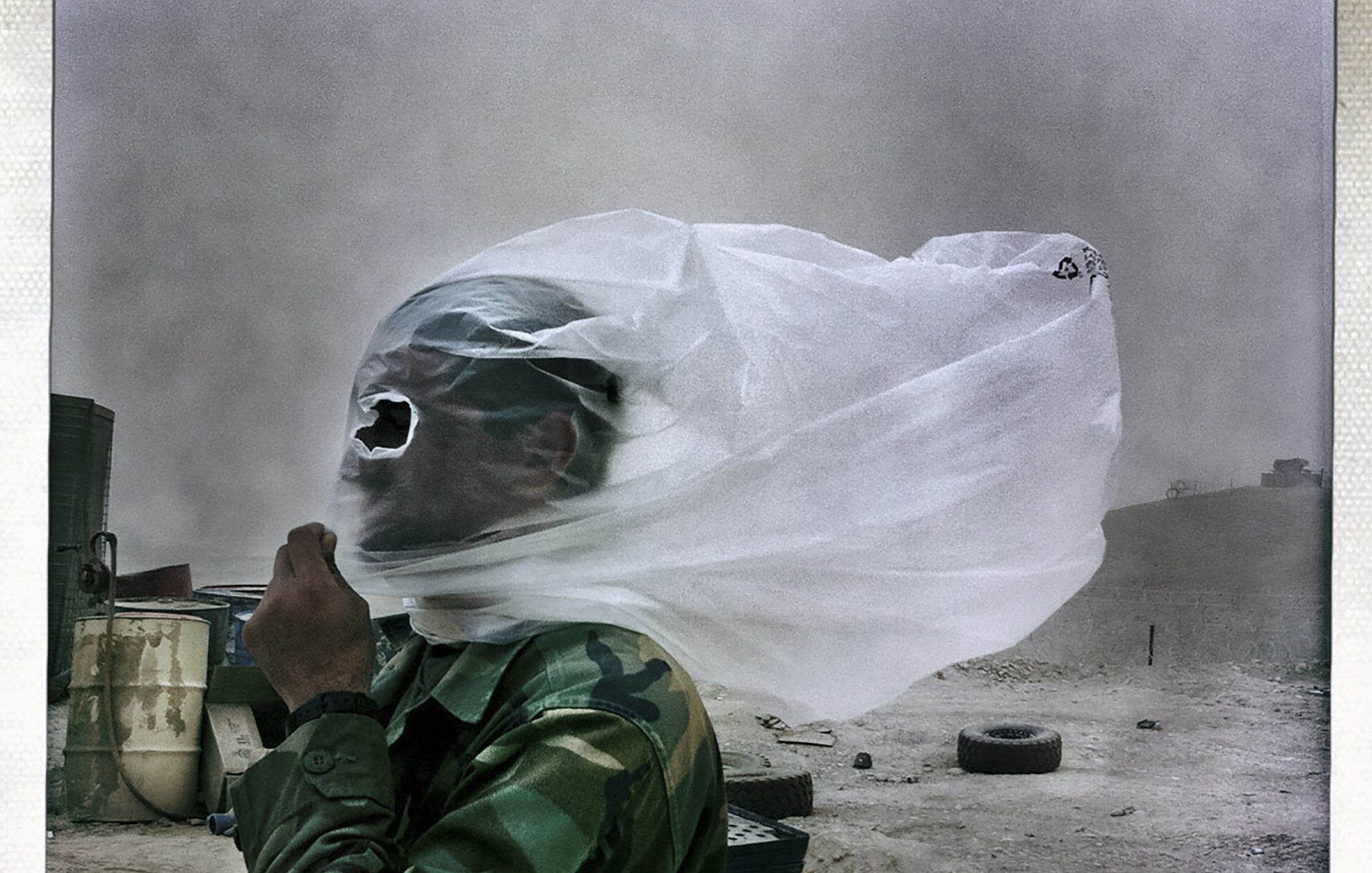 L'immagine mostra un uomo in uniforme militare, avvolto da un velo di stoffa che sembra fluttuare nel vento. Il suo viso è parzialmente coperto, rendendolo quasi irriconoscibile. Sullo sfondo si intravede un paesaggio desolato, con sabbia e detriti, suggerendo che si trovi in un ambiente arido, probabilmente in una zona di conflitto. L'atmosfera appare drammatica a causa del vento che solleva la polvere.