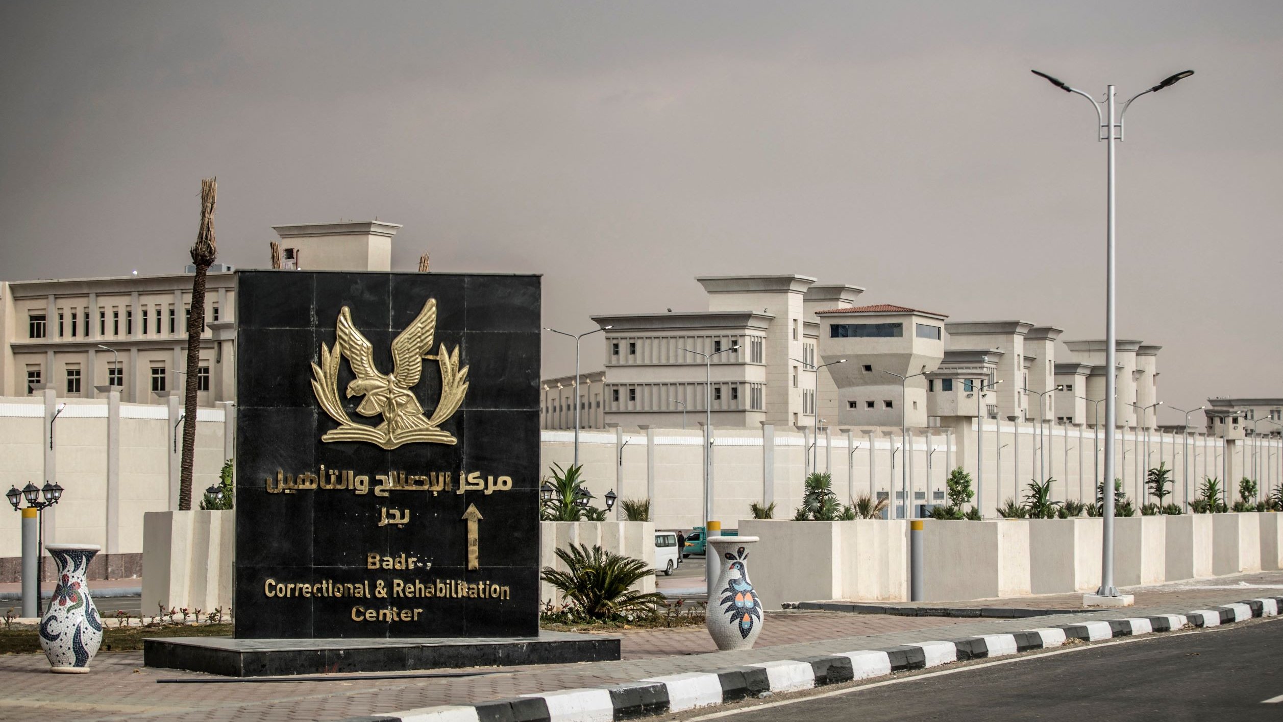L'immagine mostra un grande cartello di benvenuto che indica il "Centro di Correzione e Riabilitazione di Badia". Il cartello è di colore scuro con un simbolo o un emblema, e si trova all'ingresso di una struttura recintata. Sullo sfondo si possono vedere edifici moderni, circondati da piante e un'illuminazione stradale. Il contesto appare ordinato e ben tenuto, suggerendo un'area istituzionale.