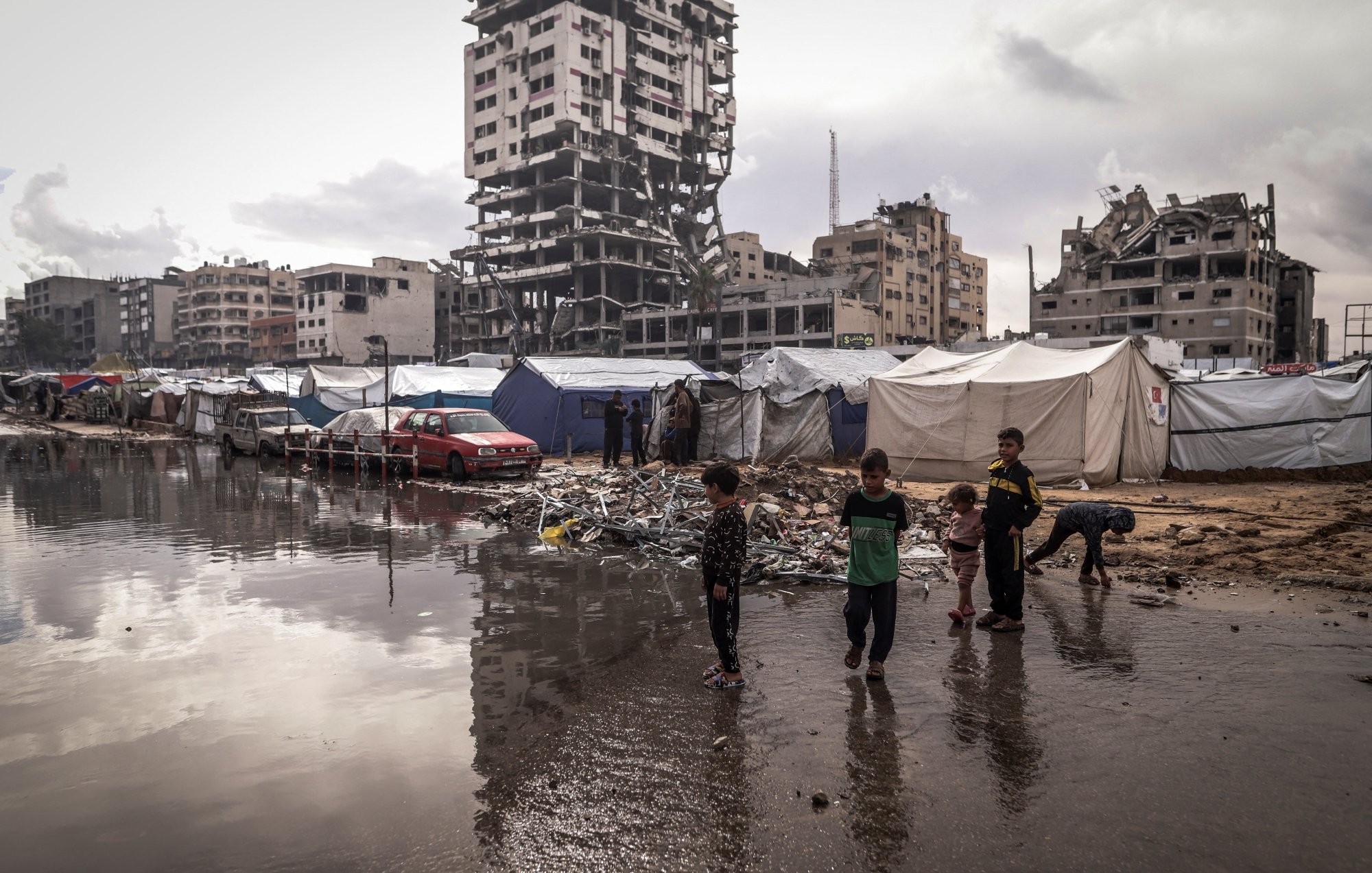 Un campo profughi con tende, bambini giocano e un edificio in rovina sullo sfondo.
