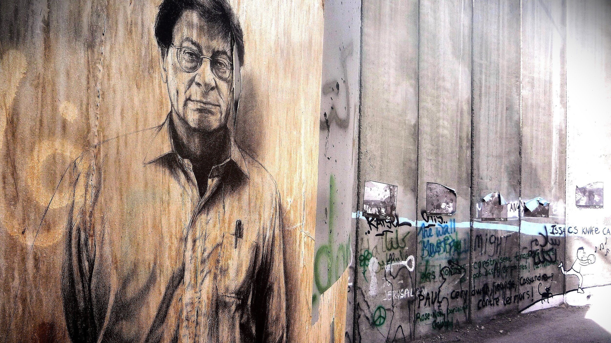 L'immagine rappresenta un murale su un muro di cemento, caratterizzato da un ritratto di un uomo con occhiali e un'espressione seria. Il muro è grezzo e presenta anche scritte e graffiti, suggerendo un contesto urbano e di protesta. Il contrasto tra il ritratto e il resto del muro evidenzia un messaggio artistico e sociale. La scena evoca un senso di riflessione e di critica.