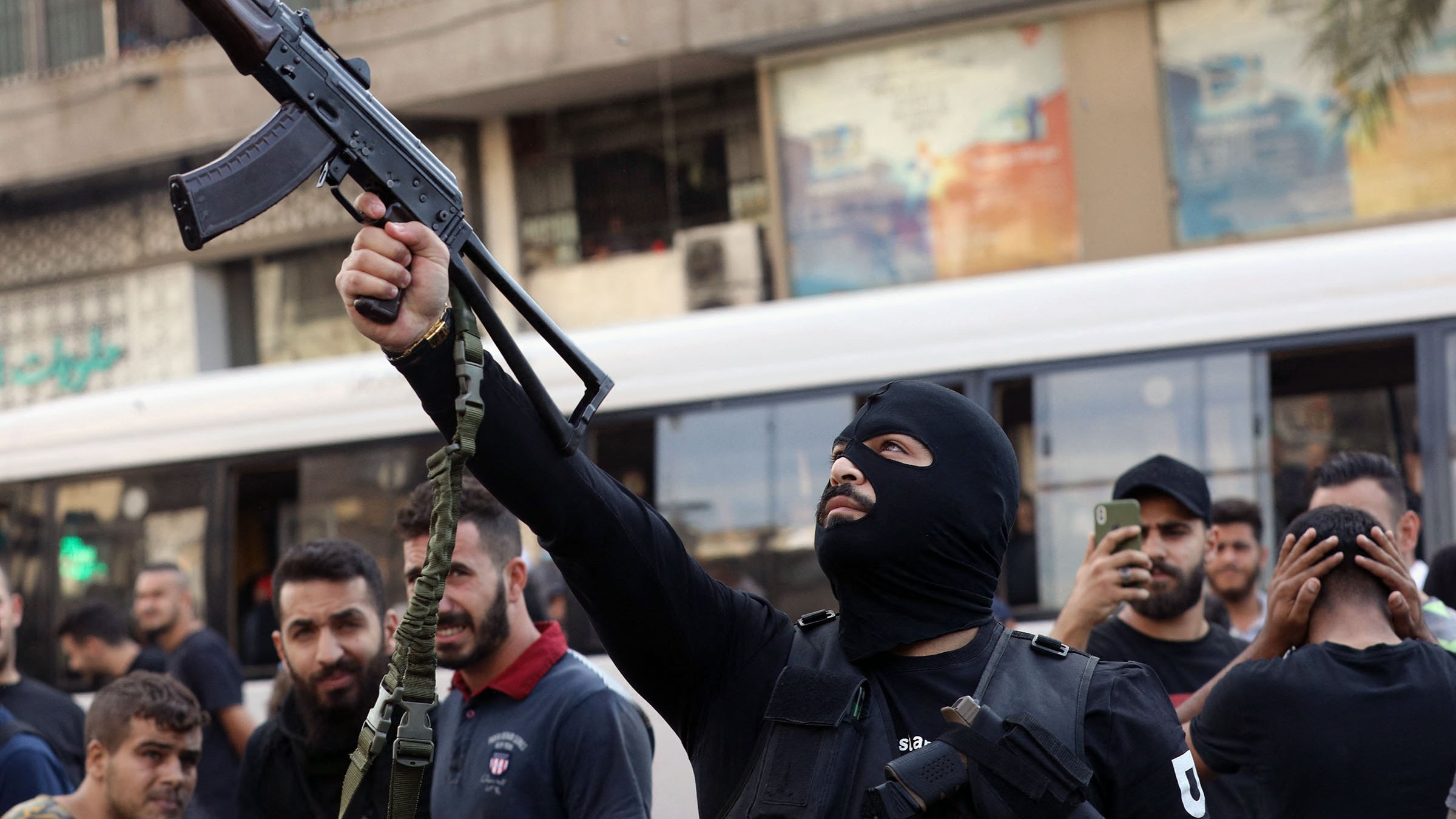 L'immagine mostra un uomo mascherato che brandisce un fucile automatico in una strada affollata. Si possono vedere altre persone intorno a lui, alcune delle quali sembrano sostenere o osservare l'azione. L'ambiente circostante è urbanizzato, con edifici e un autobus che appare in lontananza. L'atmosfera sembra carica di tensione, suggerendo un contesto di protesta o conflitto.