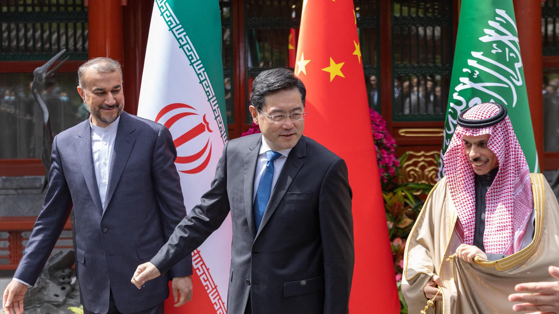 L'immagine mostra tre uomini che si trovano in un contesto formale e diplomatico, con bandiere dell'Iran, della Cina e dell'Arabia Saudita in background. I soggetti sembrano essere coinvolti in una discussione o in un incontro. L'atmosfera sembra prestigiosa e rappresenta un momento di incontro tra le nazioni.