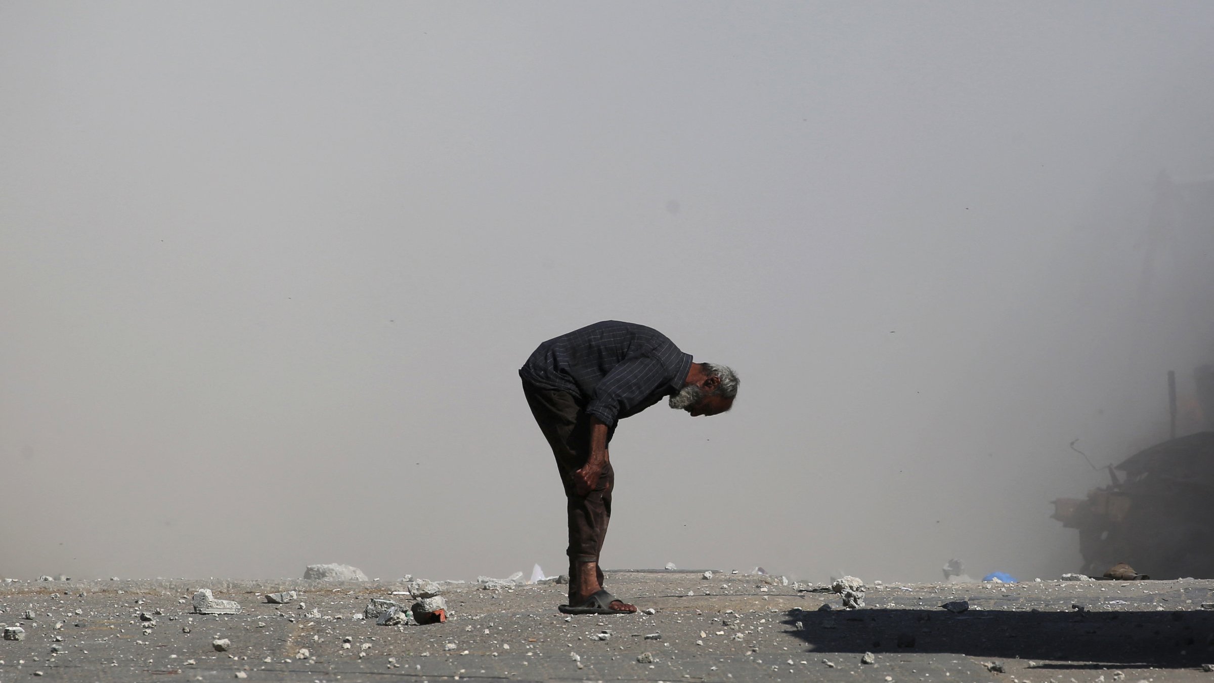 Un uomo anziano si piega in avanti su una strada polverosa, circondato da detriti.