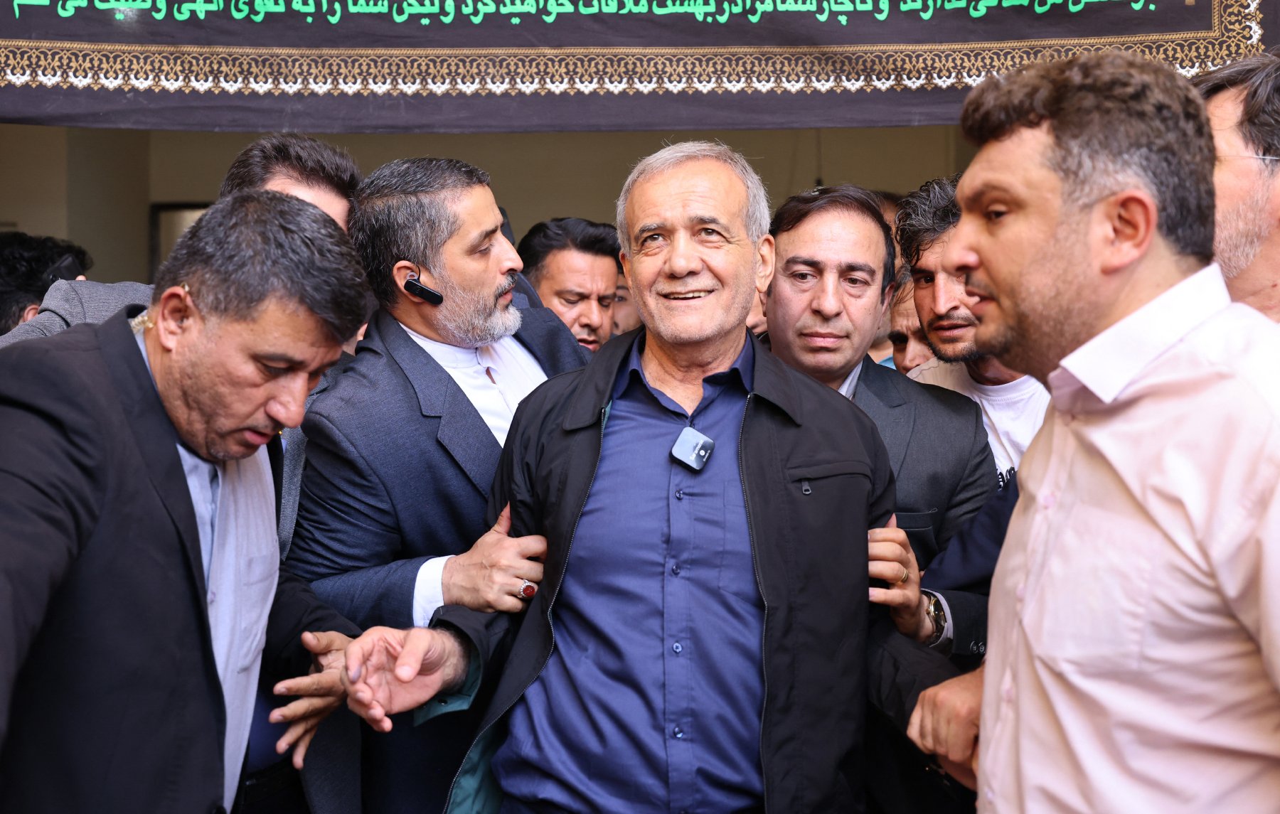 L'immagine mostra un gruppo di uomini in una situazione di affollamento. Al centro, un uomo di età avanzata sembra essere il focus dell'attenzione, mentre gli altri uomini lo circondano, alcuni di loro lo toccano o lo sostengono. L'atmosfera appare intensa e focalizzata, con uno sfondo decorato da un drappo o un banner che contiene testo in un'altra lingua. L'espressione dell'uomo centrale è di soddisfazione o di riconoscenza.