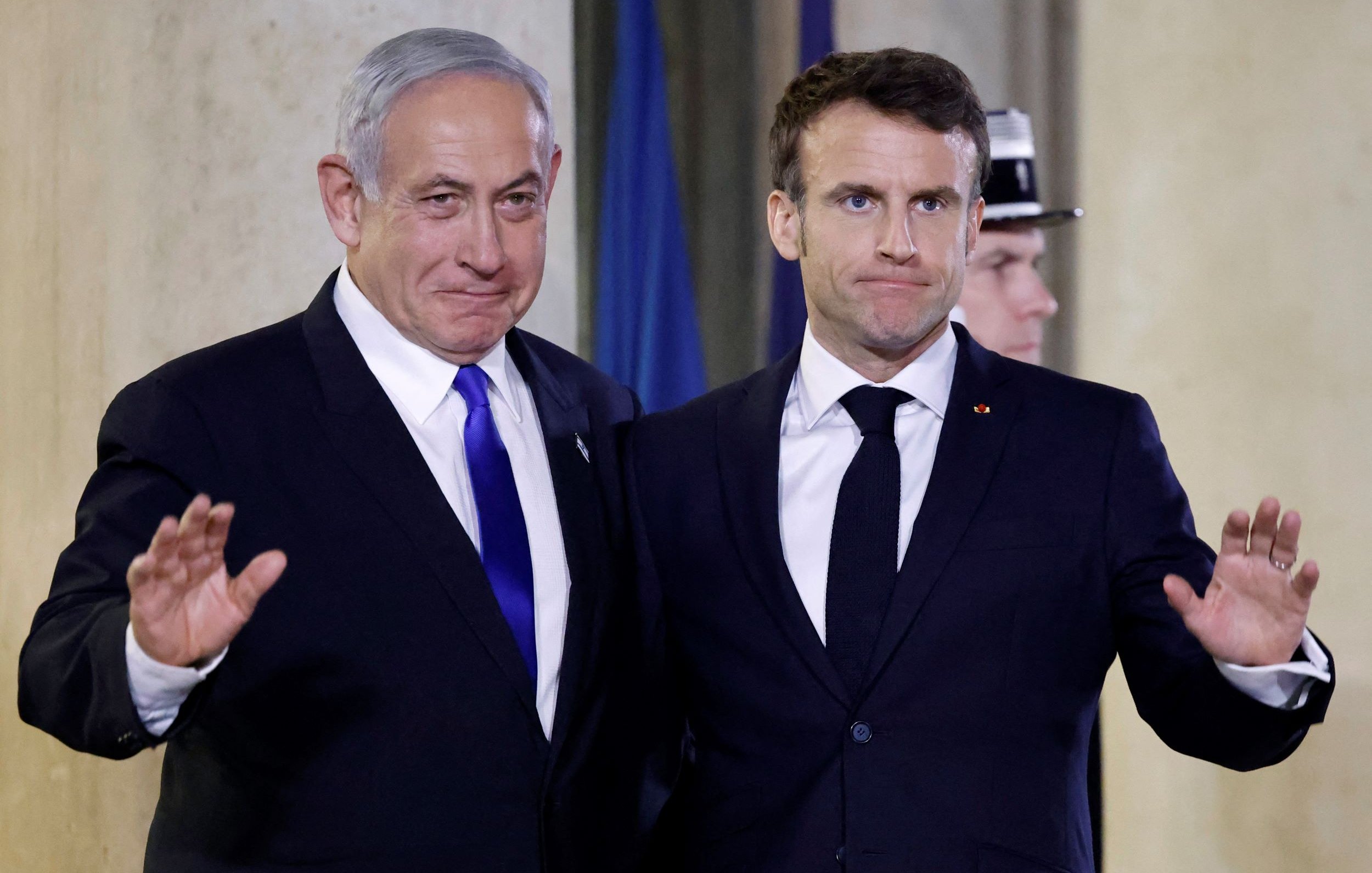 Nell'immagine si vedono due uomini in abiti formali, sorridenti e in posa per una foto. Sono entrambi in piedi, con una mano sollevata in segno di saluto. Lo sfondo presenta un ambiente elegante, probabilmente all'interno di un edificio istituzionale. Gli uomini sembrano essere in un contesto ufficiale, come una cerimonia o un incontro diplomatico.