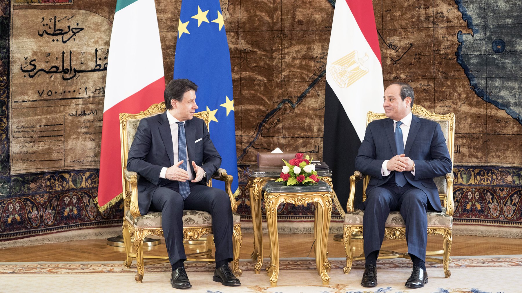 L'immagine mostra due uomini seduti su poltrone dorate in un ambiente elegante. Sullo sfondo si possono notare le bandiere dell'Italia e dell'Egitto, insieme a una bandiera dell'Unione Europea. Entrambi gli uomini stanno conversando e sembrano impegnati in una discussione formale, circondati da elementi decorativi e un tappeto elaborato. Sulle loro ginocchia ci sono bouquet floreali. L'atmosfera è di cordialità e ufficialità.
